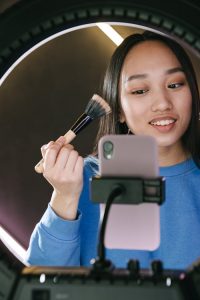 Young woman streaming a beauty tutorial using a ring light and mobile phone, applying makeup indoors.